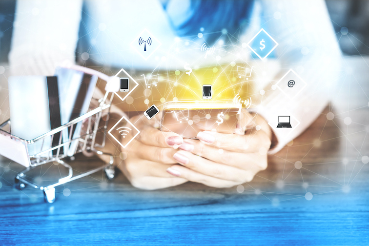 A shopper holds a smartphone beside a small shopping cart, with digital shopping and connectivity icons over the image.