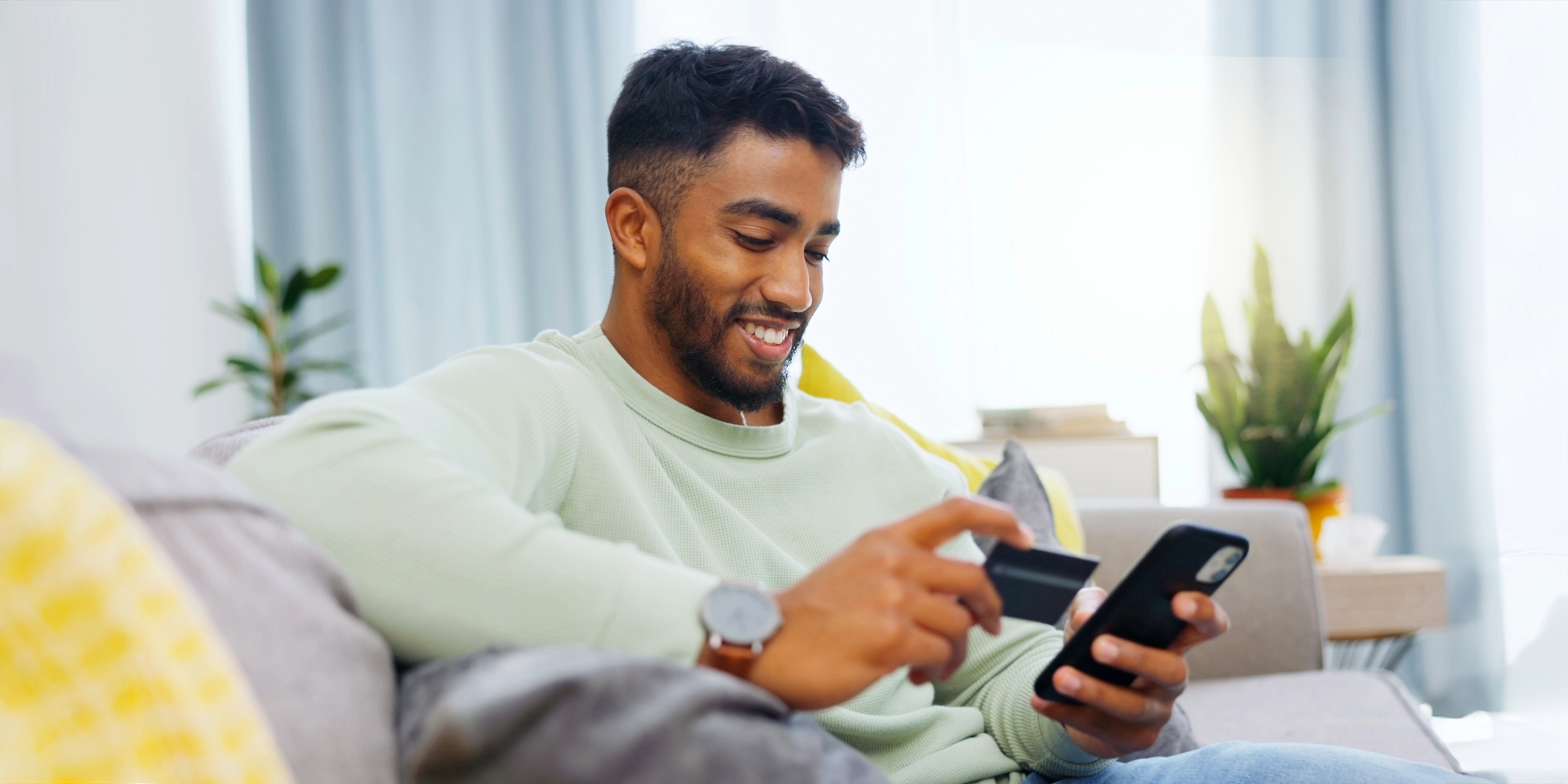 A man sits on a couch smiling as he shops with a smartphone and credit card.